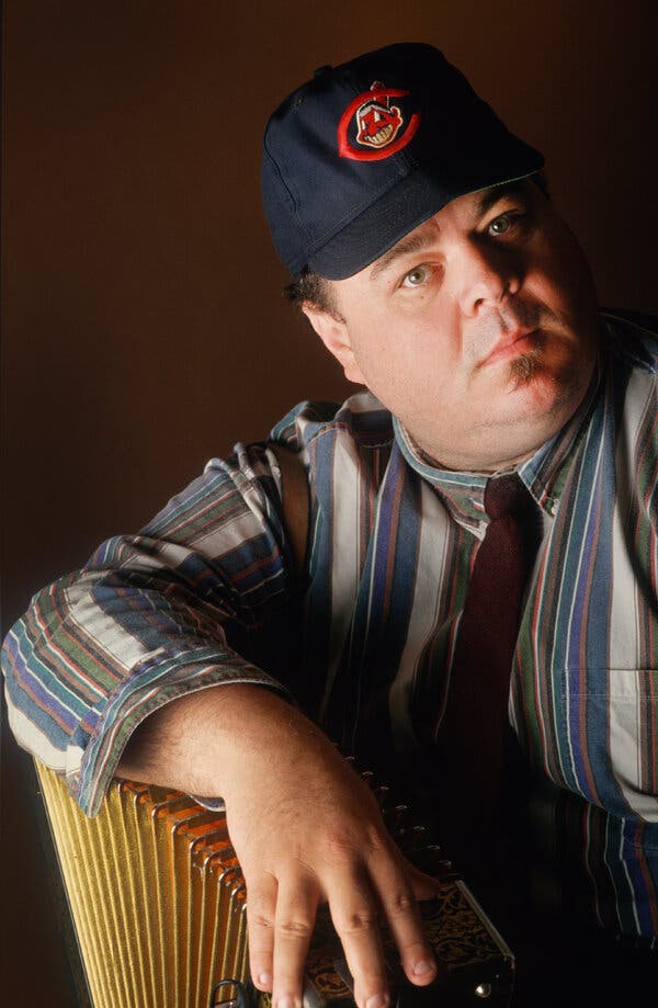 In a close-up photo, he wears a Cleveland Indians baseball cap, a loud striped shirt and a necktie and holds an accordion. 