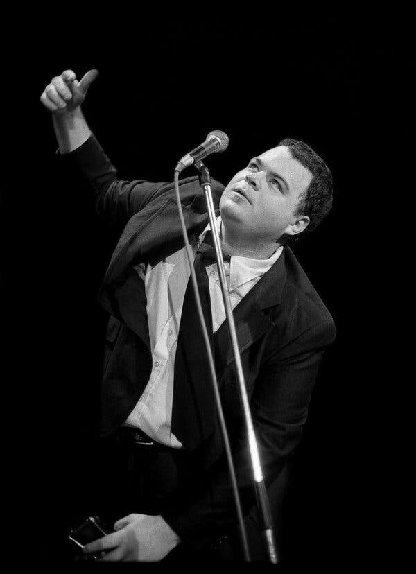 A black and white photo of him onstage in an unbuttoned dark suit, white shirt and dark tie, looking up as he stands before a microphone with his right arm raised. 
