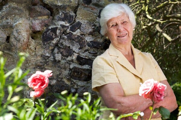 Jane Gardam, who has white hair and is wearing a yellow shirt, leans against a tree. In front of her are pink roses.