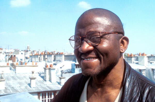 A close-up photo of him smiling for the camera on a rooftop in Paris, the rooftops of other buildings spread out behind him. 