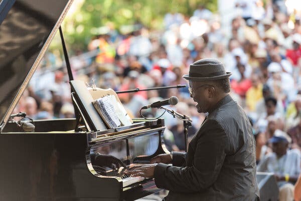Wearing a porkpie hat, he sits at a grand piano onstage singing into a microphone as he plays the instrument before a large crowd. 